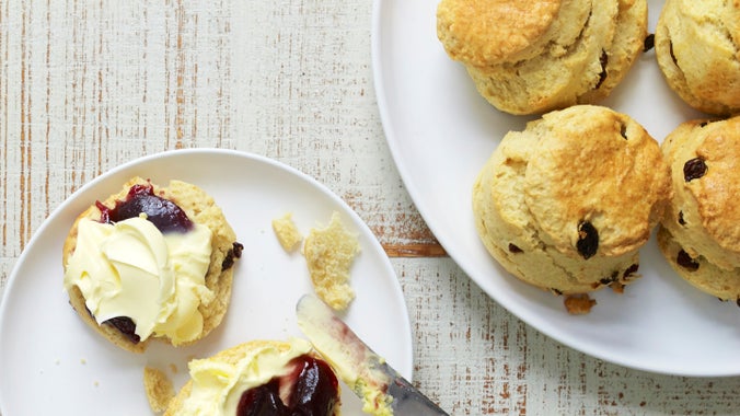 A halved fruit scone on a plate topped with jam and clotted cream, and a plate of fruit scones